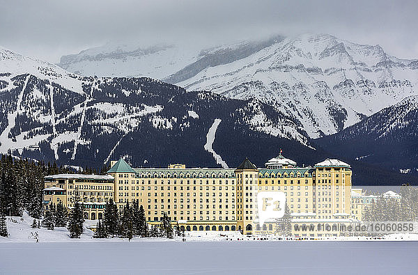 Large Chateau Hotel lit by the sun with a mountain ski hill in the background and snow-covered Lake Louise in the foreground  Banff National Park; Lake Louise  Alberta  Canada