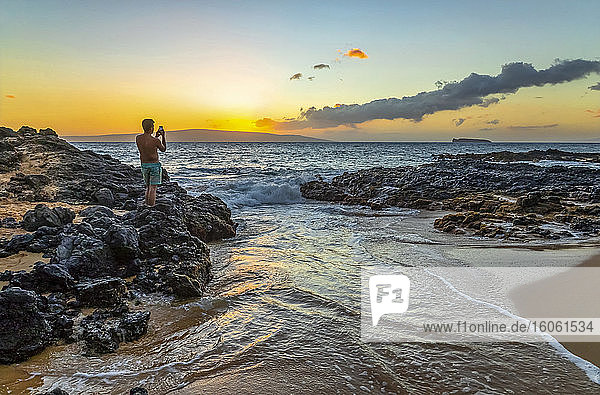 A young man in a bathing suit stands on the volcanic rock on the shore capturing the golden sunset with a smart phone camera; Kihei  Maui  Hawaii  United States of America