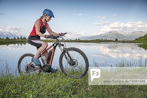 Mountain biker  in her late forties  rides an e-bike along the lake shore against a mountain backdrop  Mutterer Alm  Stubaier Alpen  Mutters  Tyrol  Austria  Europe