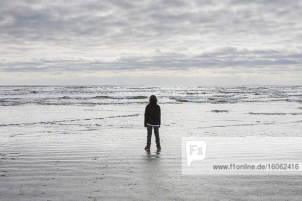 Teenager Junge steht auf riesigen Strandwellen und bedecktem Himmel in der Ferne