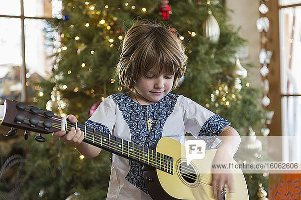 lächelnder 4-jähriger Junge spielt Gitarre mit Weihnachtsbaum im Hintergrund