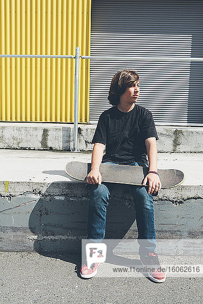 Teenage boy posing siting with skateboard in front urban warehouse