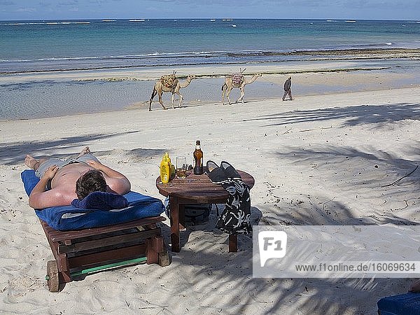 Touristen sonnen sich in einer Ferienanlage  während im Hintergrund ein Einheimischer Kamele am Strand von Tiwi an der Küste des Indischen Ozeans  Kenia  zieht.