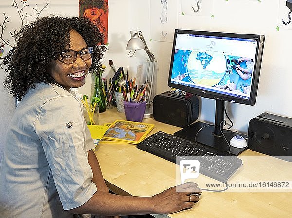 Dark-skinned  smiling woman with glasses works at her desk in front of a computer  home office  looking into the camera  Germany  Europe