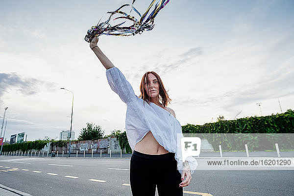 Portrait of woman with long red hair  standing on street  holding streamers in raised hand.