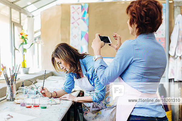 Künstlerin malt im Kreativstudio auf Stoff  reife Frau fotografiert sie mit dem Smartphone