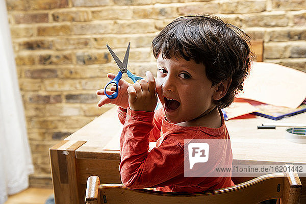 Junge Junge mit braunen Haaren sitzt am Tisch  hält eine Schere in der Hand und lächelt in die Kamera.