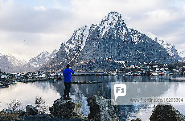 Rear view of male tourist photographing snow capped mountain and village waterfront  Reine  Lofoten  Norway