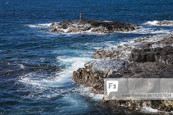 Schwarze Lavafelsen an einer zerklüfteten Küstenlinie mit brechenden Wellen  eine Person steht auf einem Felsvorsprung.