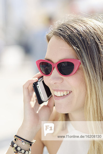 Kopfschuss einer lächelnden jungen Frau mit Sonnenbrille  die ihr Mobiltelefon im Freien benutzt  Portugal  Europa