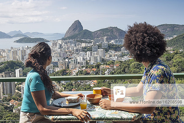 Ein multiethnisches Ehepaar frühstückt gemeinsam und blickt auf den Zuckerhut und die Skyline von Rio  Rio de Janeiro  Brasilien  Südamerika
