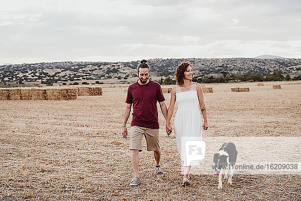 Lächelndes Paar hält sich an den Händen  während es mit seinem Hund in einem Feld spazieren geht