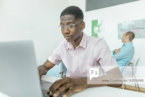 Ein Mitarbeiter arbeitet an einem Laptop  während ein Kollege im Hintergrund am Arbeitsplatz sitzt