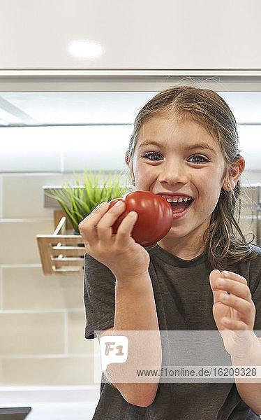 Nahaufnahme eines fröhlichen Mädchens  das eine Tomate in der Hand hält  während es in der Küche zu Hause sitzt