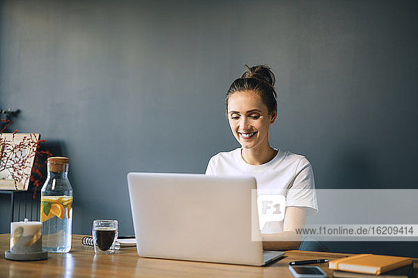Lächelnde Geschäftsfrau mit Laptop auf dem Schreibtisch an der Wand im Heimbüro
