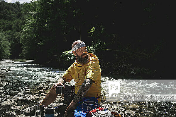Wanderer mit Vollbart und gelbem Kapuzenpulli bei der Pause  Tee kochen am Flussufer