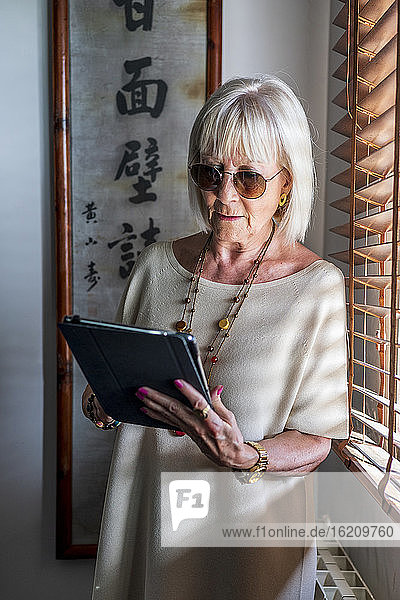 Ältere Frau mit Sonnenbrille  die ein digitales Tablet benutzt  während sie zu Hause am Fenster steht