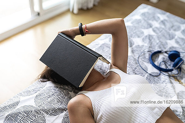 Mittlere erwachsene Frau mit Buch auf dem Gesicht entspannt im Schlafzimmer zu Hause