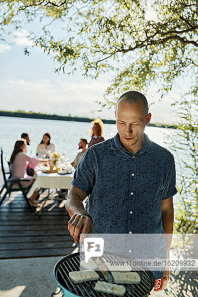 Man grilling Halloumi cheese at a lake with friends in background