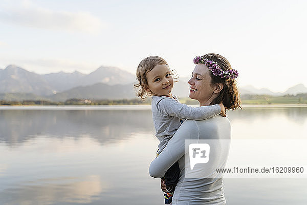 Lächelnde Mutter mit Diadem  die ihre Tochter trägt  während sie bei Sonnenuntergang am See steht