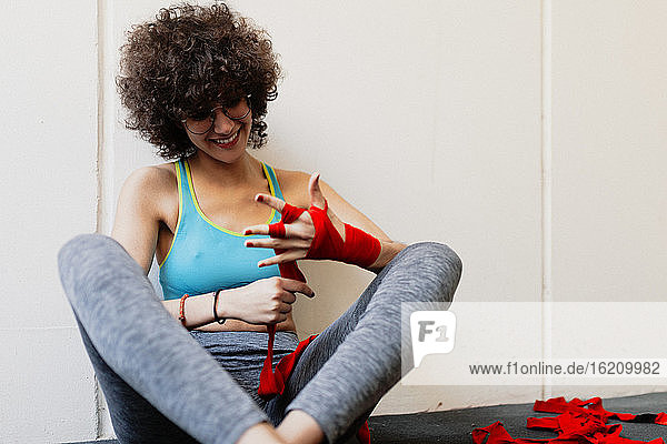 Weiblicher Boxer mit Afro-Haar wickelt roten Verband um die Hand  während er im Fitnessstudio sitzt