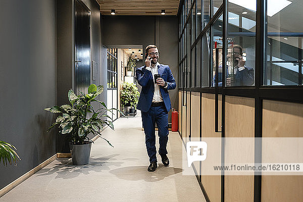 Lächelnder Geschäftsmann  der mit seinem Handy telefoniert  während er eine Tasse an einer Glaswand im Bürokorridor vorbeischiebt