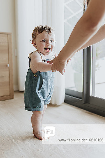 Ein kleines Mädchen hält die Hände seiner Mutter und lernt zu Hause auf dem Boden zu laufen