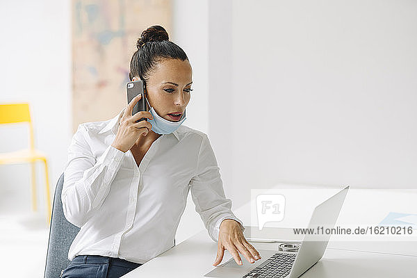 Geschäftsfrau mit Maske  die über ein Smartphone spricht  während sie einen Laptop auf dem Schreibtisch im Heimbüro benutzt