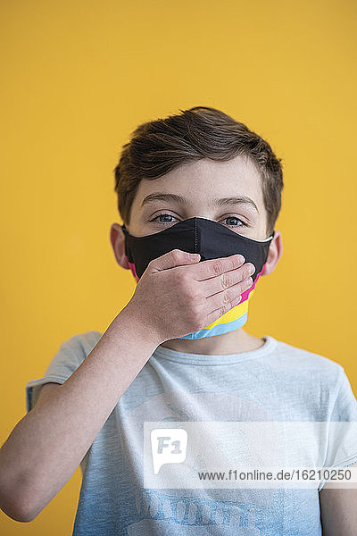 Close-up of boy wearing mask covering mouth with hand against yellow background