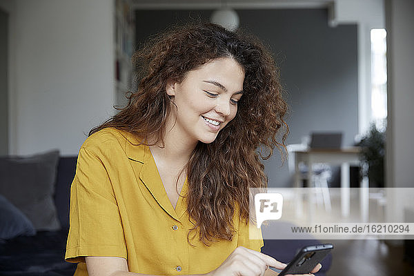 Junge Frau lächelt  während sie zu Hause eine Textnachricht auf ihrem Smartphone schreibt