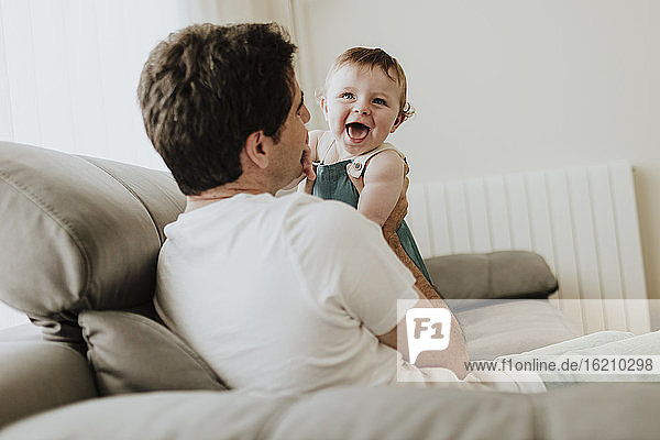 Father holding cheerful baby daughter while relaxing on sofa at home