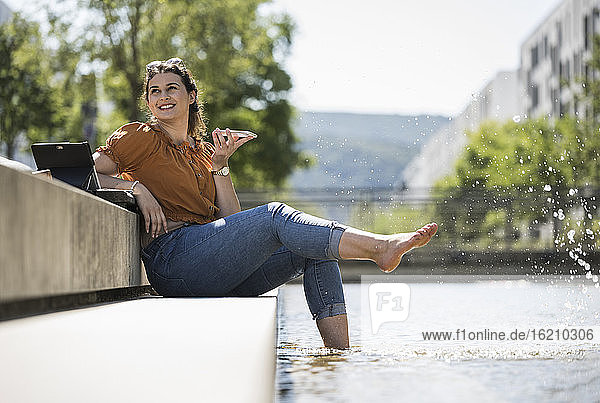 Junge Frau mit Beinen im Teich  die über ihr Smartphone spricht  während sie im Park sitzt