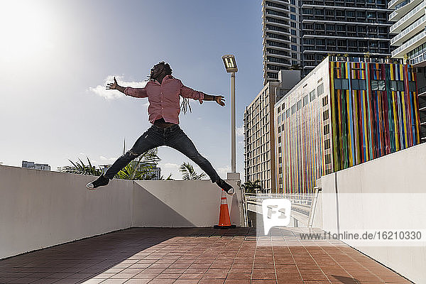 Fröhlicher Afro-Mann mit ausgestreckten Armen  der auf dem Fußweg gegen den Himmel springt  Miami  Florida  USA