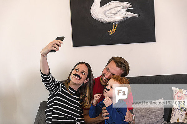 Frau mit künstlichem Schnurrbart macht Selfie mit Familie auf Sofa zu Hause