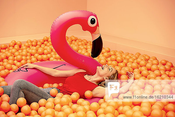 Fröhliche junge Frau mit aufblasbarem Flamingo  der in einem orangefarbenen Bällebad liegt