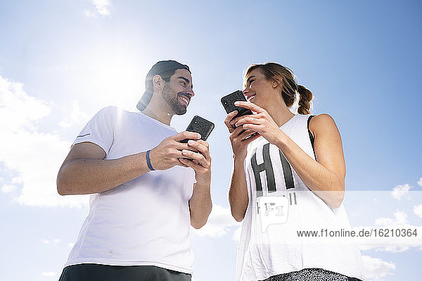 Smiling couple holding smart phones looking at each other while standing against sky during sunny day