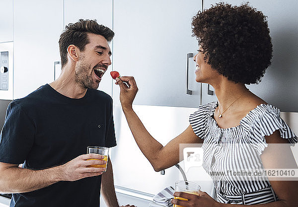 Happy woman feeding strawberry to man in kitchen of penthouse