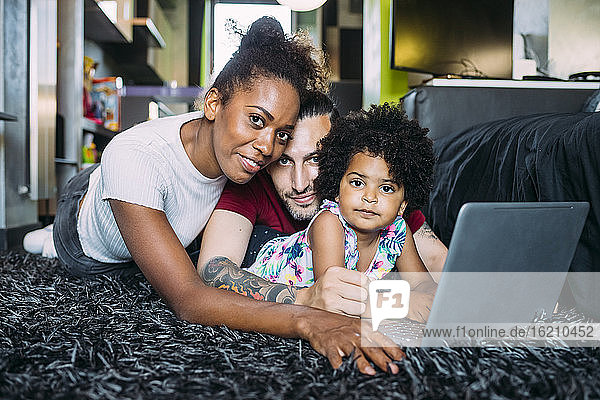 Eltern mit Tochter  die einen Laptop benutzt  während sie zu Hause auf dem Teppich liegt