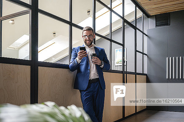 Smiling businessman holding disposable coffee cup while using smart phone against glass wall at coworking office