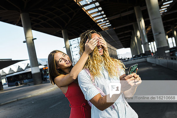 Junge Frau bedeckt die Augen ihres Hipster-Freundes von hinten  während sie auf der Straße in der Stadt steht