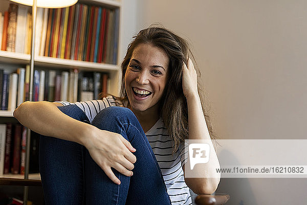 Cheerful young woman with hand in hair sitting against wall at home