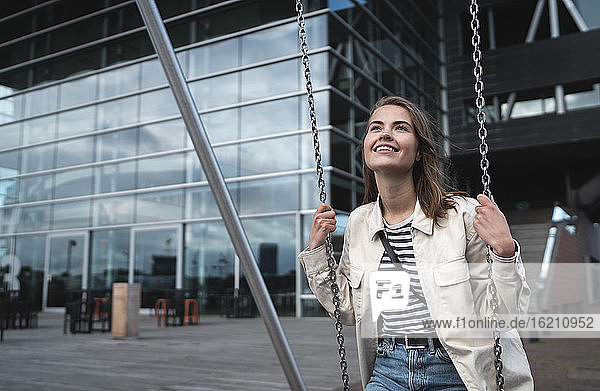Lächelnde junge Frau sitzt auf einer Schaukel vor einem modernen Gebäude in der Stadt