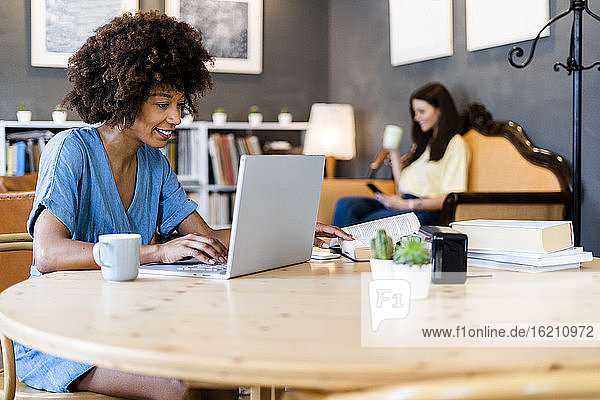 Glückliche Frau mit Laptop in einem Café mit einem Freund im Hintergrund