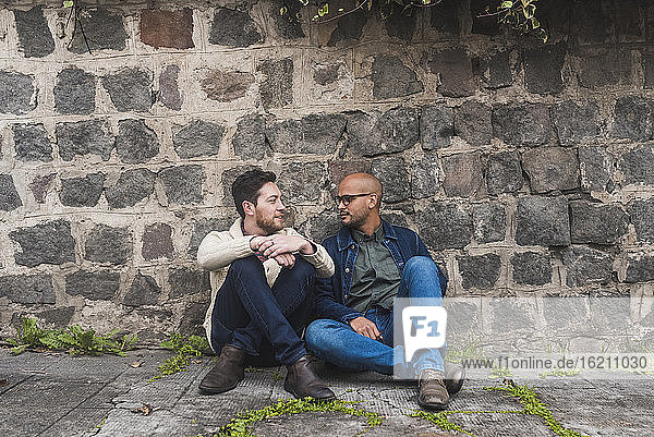 Gay couple talking while sitting on footpath against stone wall