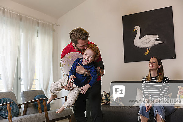 Happy woman looking at man carrying son with costume wings in living room
