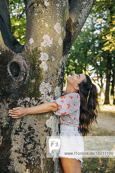 Fröhliche junge Frau mit langen Haaren  die einen Baumstamm umarmt  während sie im Park steht