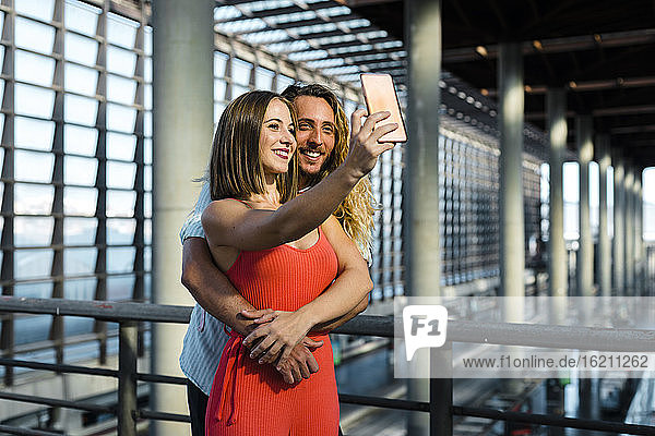 Lächelnde junge Frau  die ein Selfie mit ihrem Hipster-Freund macht  während sie am Geländer des Bahnhofs steht