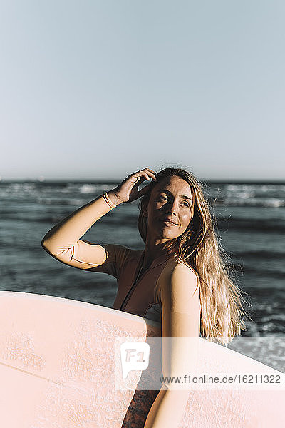 Young woman carrying surfboard looking away while standing against sea