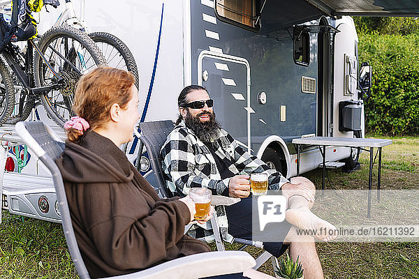 Relaxed couple sitting next to camper enjoying vacation and drinking beer