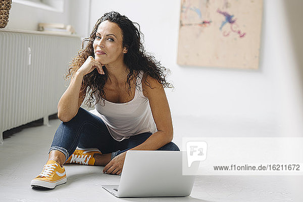 Lächelnde schöne Frau mit Hand am Kinn  die zu Hause am Laptop sitzt